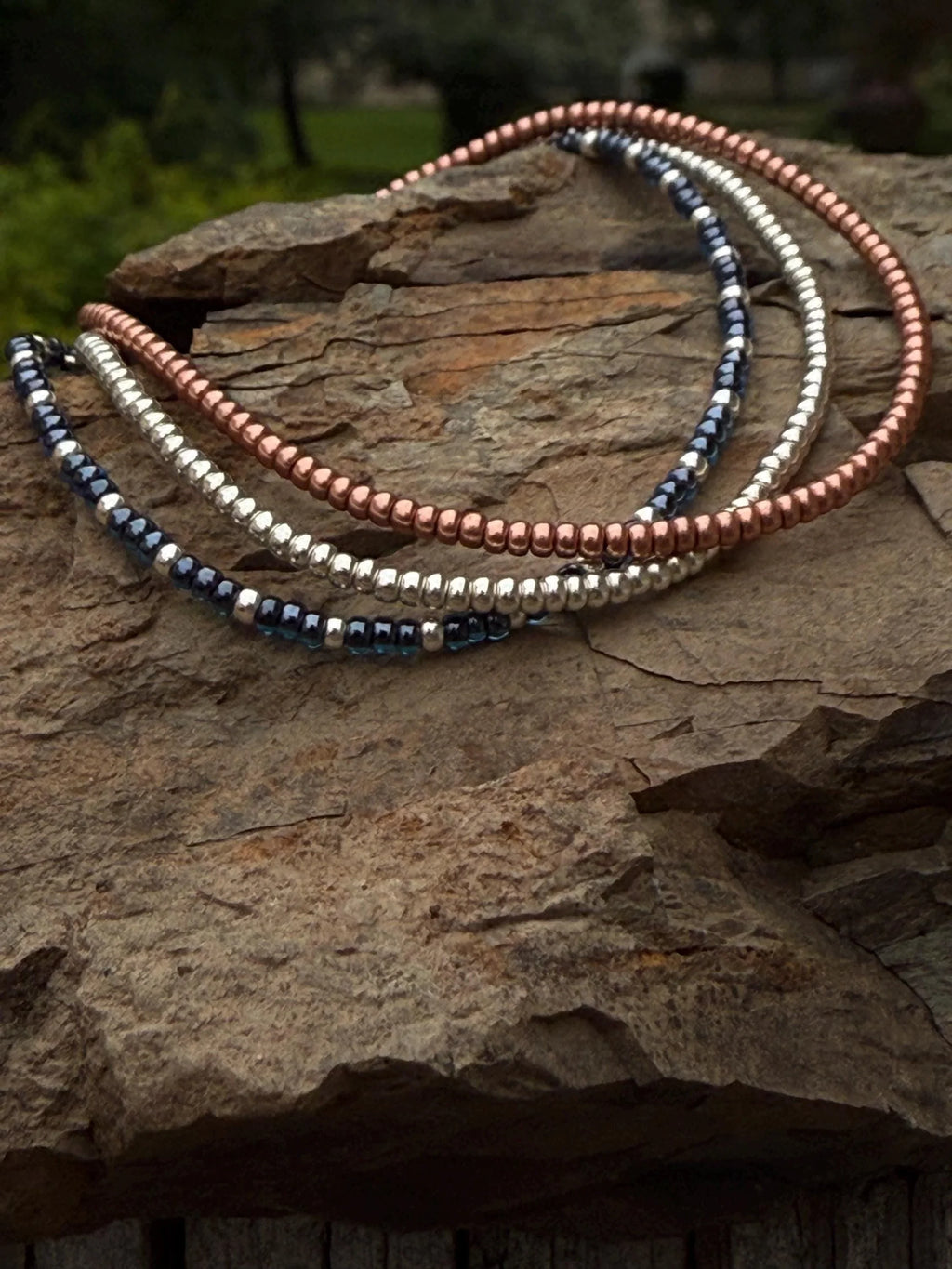 Handmade glass seed bead bracelets in metallic blue, silver, and copper on rock outdoors