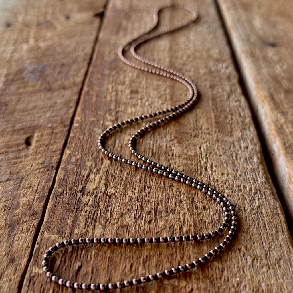 Black and copper-plated seed bead necklace laid out on a rustic wooden board.
