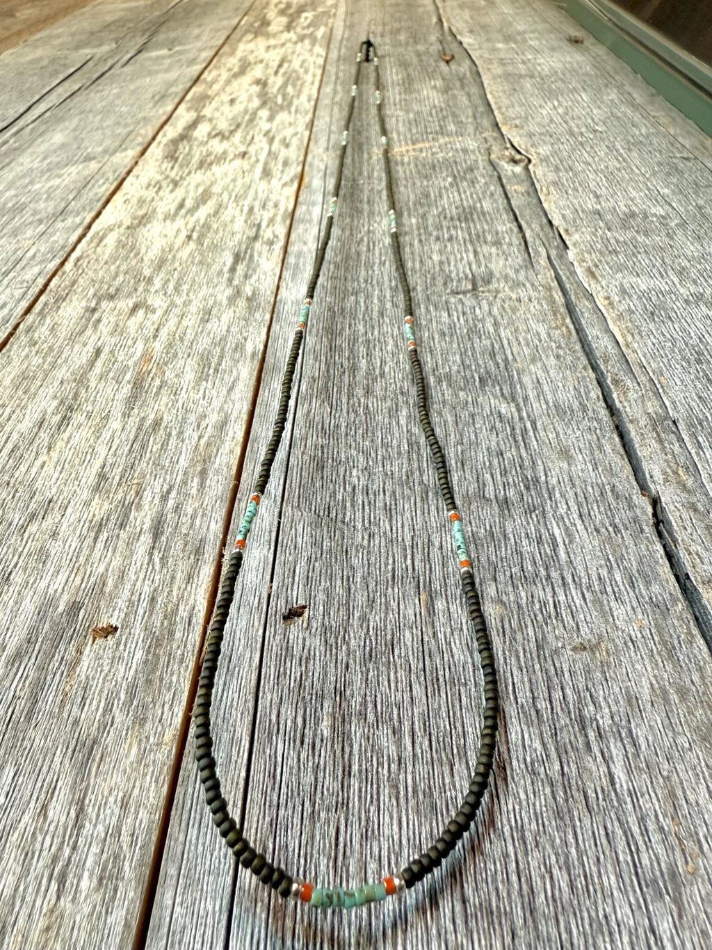 Handmade glass seed bead necklace with green and orange accents on rustic wood, Seed and Soul artistry.