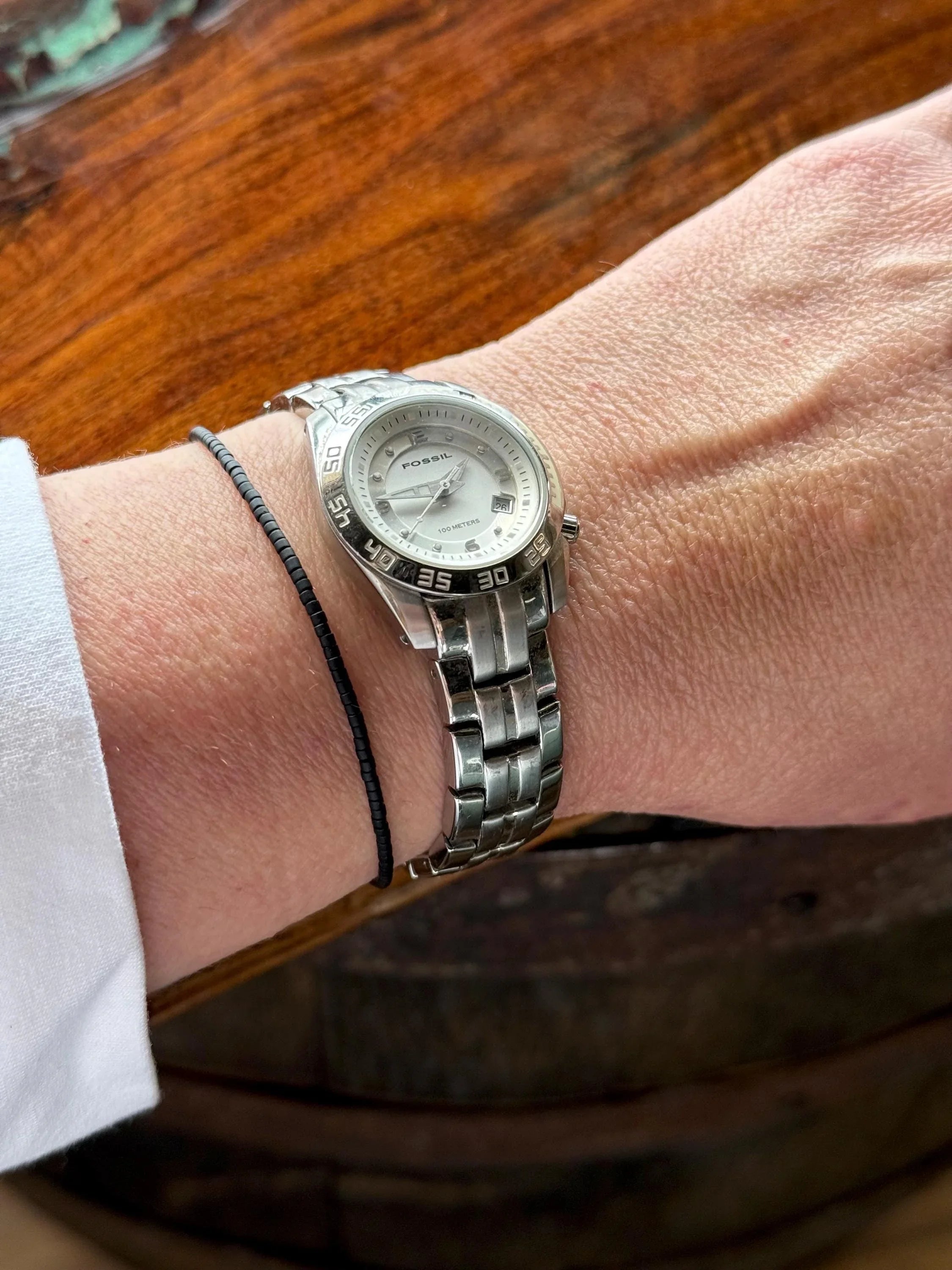 Hand wearing a thin black glass seed bead bracelet and silver watch on wood background